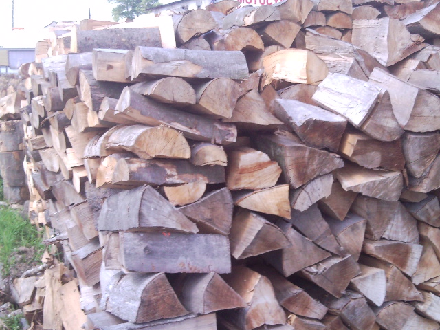 Stos pociętych kawałków drewna ułożonych na zewnątrz, widoczne fragmenty kory i sęki, w tle fragment budynku.